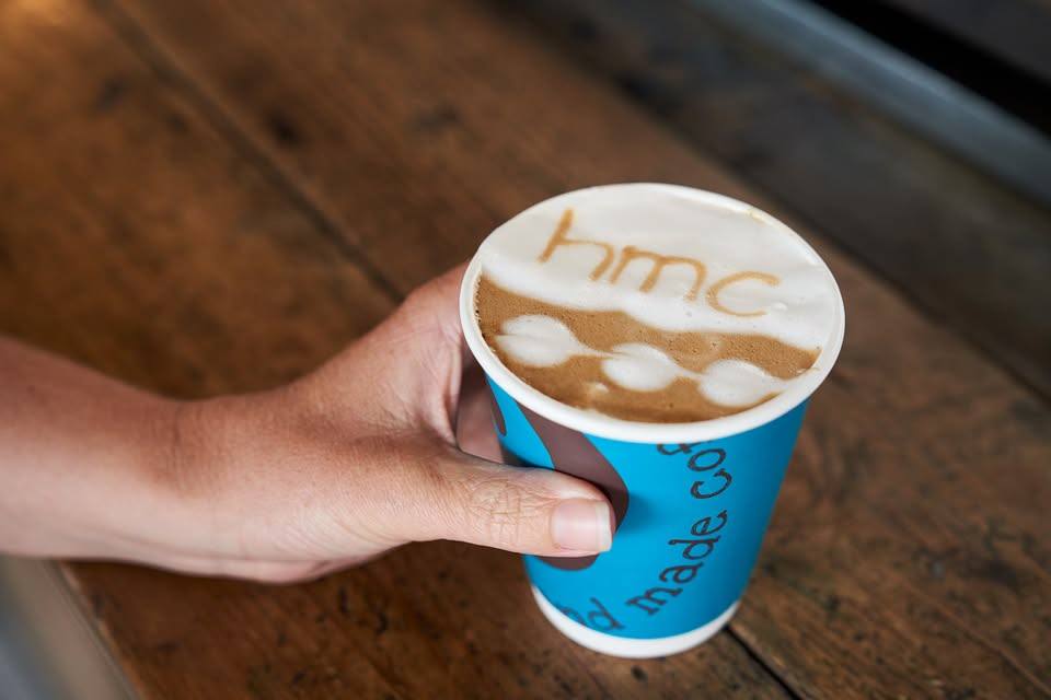A latte with “hmc” drawn in the foam, in a teal Hand Made Coffees takeaway cup, held over a worn wooden counter.