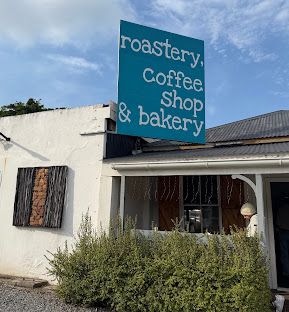 The Hand Made Coffees shopfront in Port Alfred: a whitewashed building with a teal sign reading “roastery, coffee shop & bakery” above a green veranda.